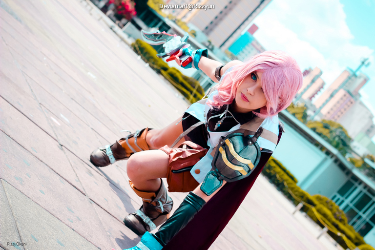 A person in a colorful cosplay outfit with pink hair, posed dramatically in an outdoor setting, holding a futuristic gun.