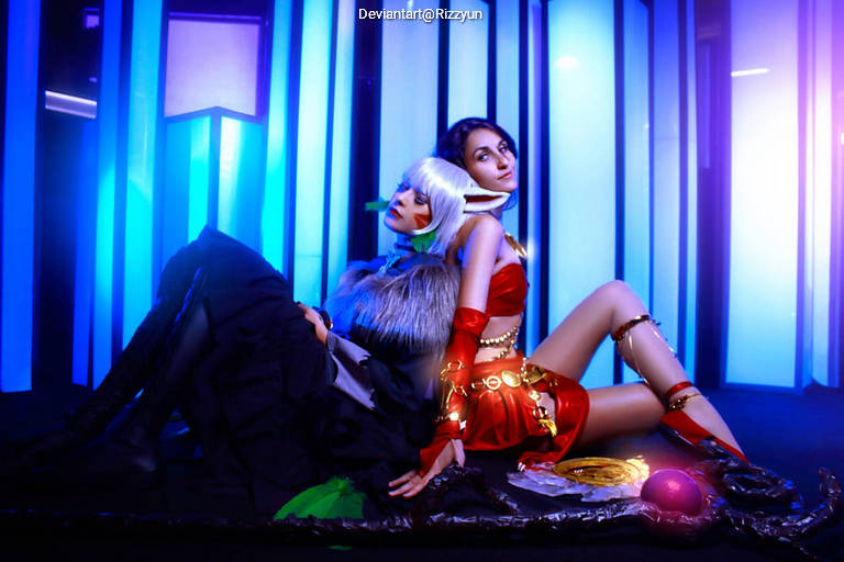 Two women in stylish costumes pose together against a backdrop of blue and purple lights, showcasing elaborate accessories and a vibrant aesthetic.