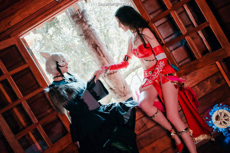 Two individuals in elaborate costumes posed indoors near a window. One person, wearing a dark outfit with animal ear accessories, is reaching toward the other, who is dressed in a vibrant red attire with intricate details. Natural light illuminates the scene.