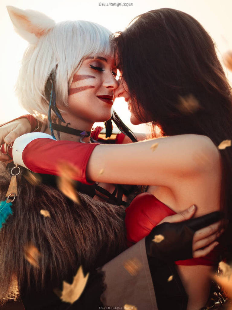 Two women embrace closely, sharing an intimate moment. One has short, white hair with animal-like features, wearing a costume with earthy tones, while the other has long, dark hair and is dressed in a red outfit. Warm tones and falling leaves create a romantic atmosphere.