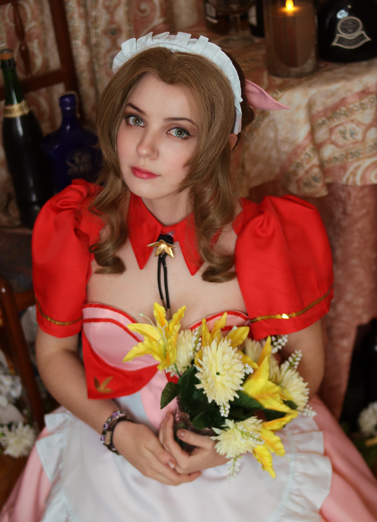 A young woman dressed in a pink and red costume with a white apron, holding a bouquet of yellow and white flowers, with a soft expression in a vintage-themed setting.