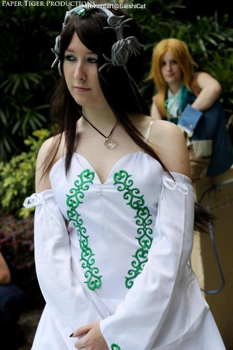 A person in a white dress with green patterns stands in a natural setting, while another person is blurred in the background.