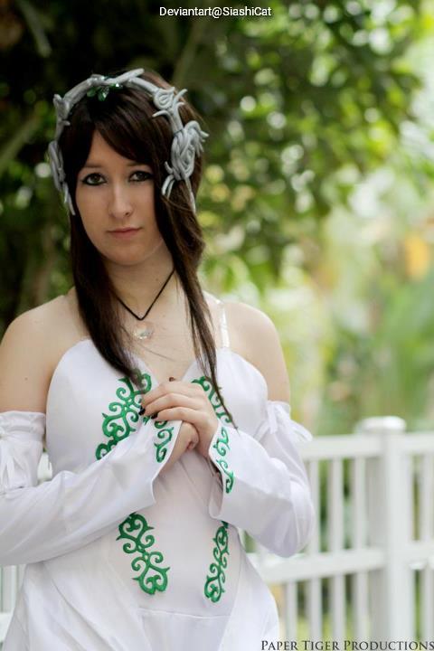A person dressed in a white costume with green designs, standing with hands clasped, amidst a blurred outdoor background.