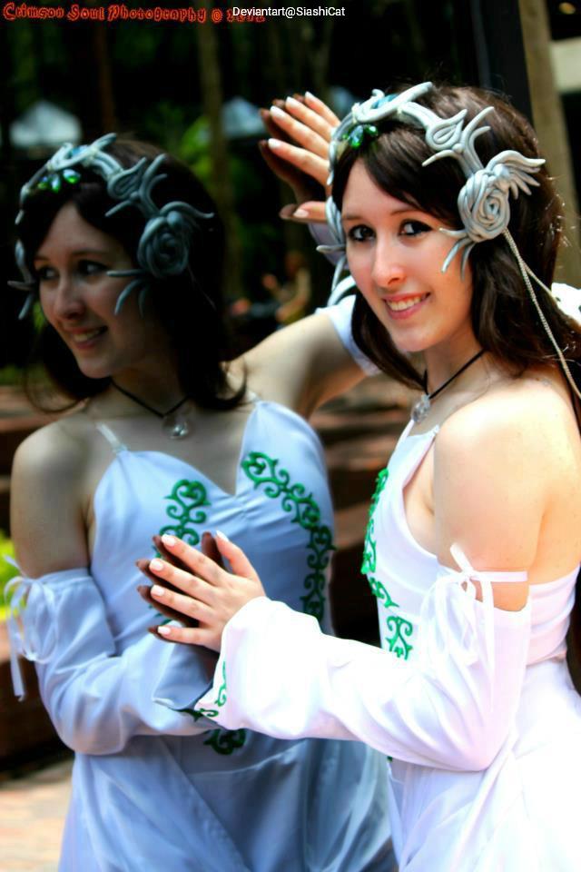 A cosplayer wearing a white dress with green accents and a floral headpiece poses in front of a mirror, smiling warmly.