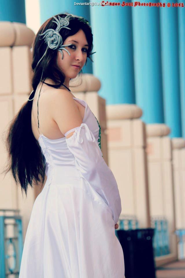 A woman with long dark hair adorned with floral accessories, wearing a white dress, poses gracefully in front of blue columns.