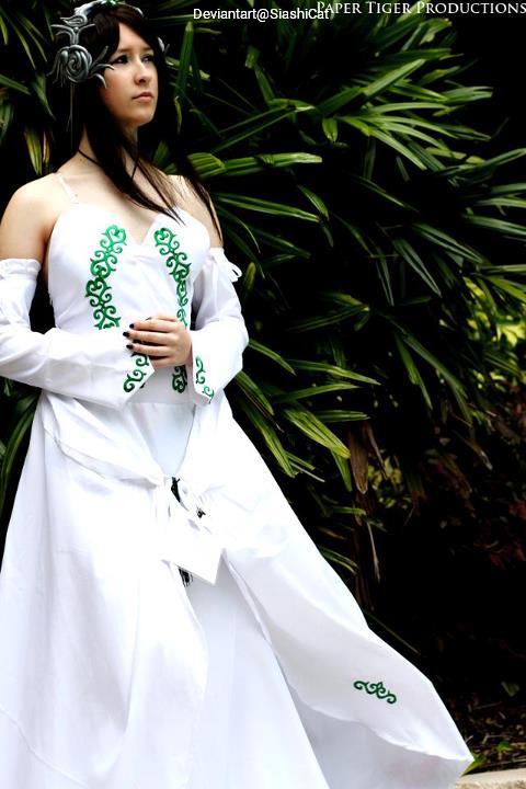 A woman in a white gown with green embroidered designs stands in front of lush greenery, looking thoughtfully into the distance.
