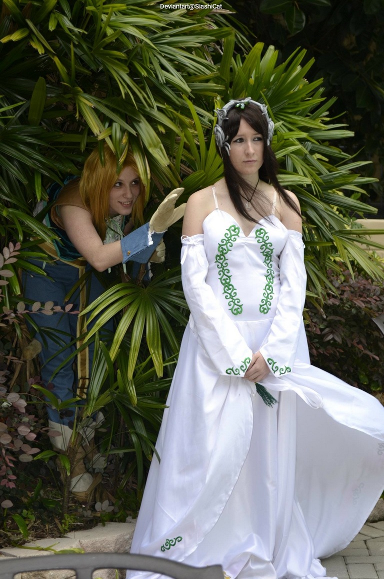 A person in a white dress with green designs stands in front of lush greenery, while another person with blonde hair peeks out from behind the plants, wearing a blue and brown outfit.