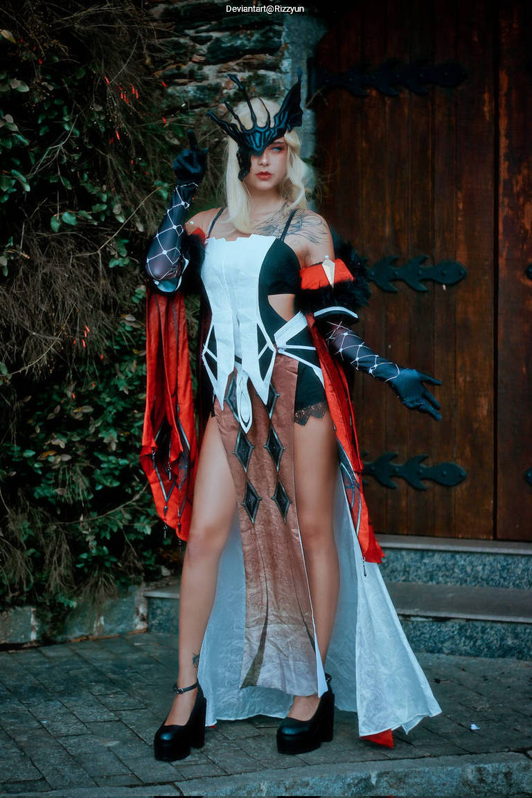 A person in an elaborate costume featuring red and black elements, standing against a wooden backdrop. The outfit includes a dramatic cape and a stylized headpiece, with high heels adding to the overall look.
