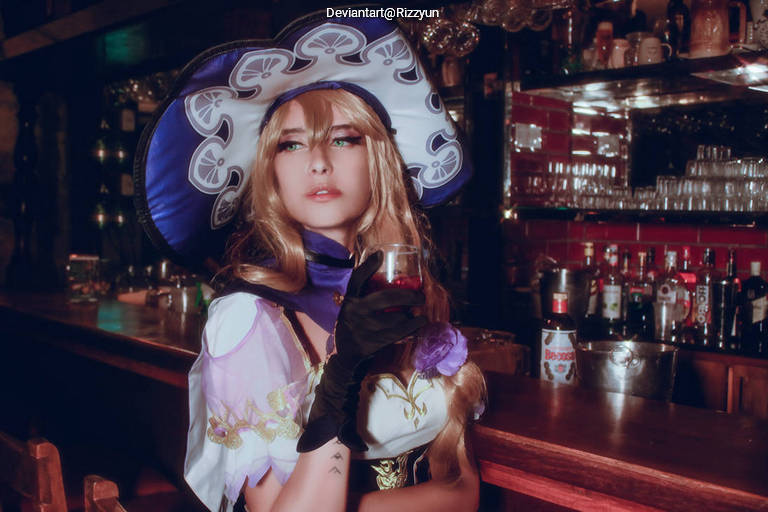 A woman in a dramatic costume with a large hat and long blonde hair holds a drink in a bar setting, with shelves of bottles in the background.