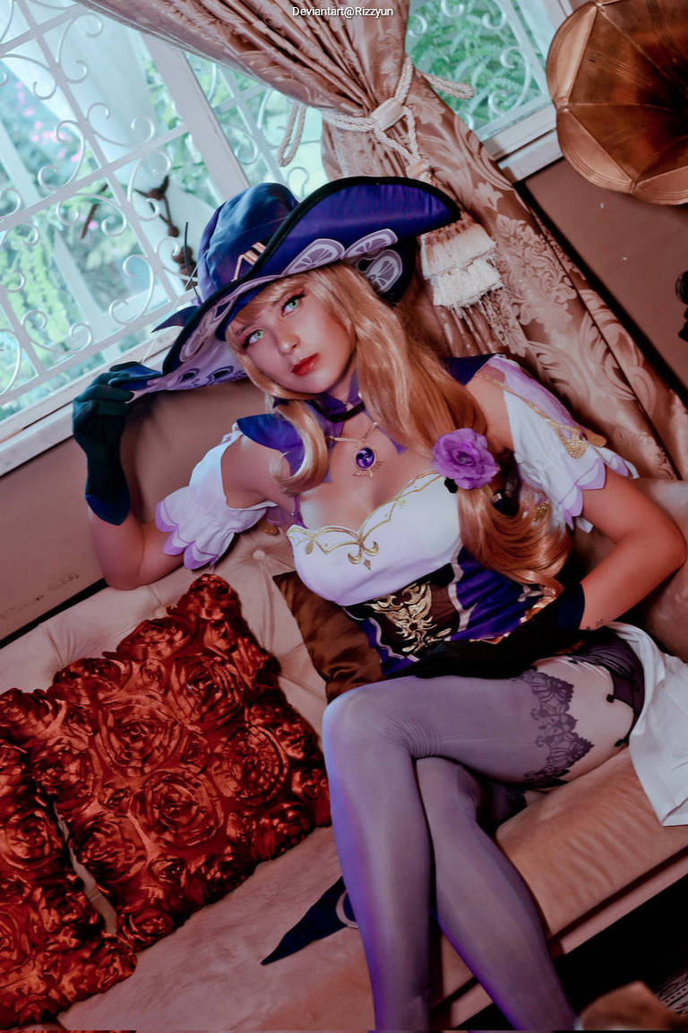 Cosplay of a character with long blonde hair, wearing a stylish blue and white outfit with a large hat, sitting on a couch surrounded by decorative pillows.