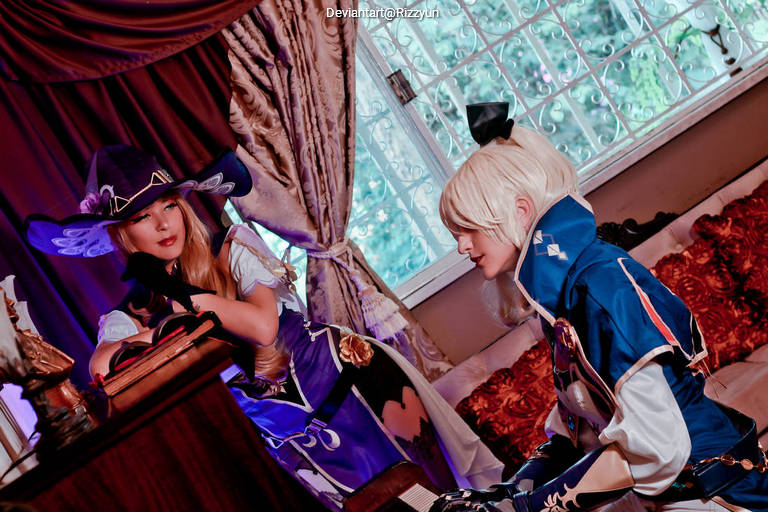 Two cosplayers in an indoor setting, one dressed as a character with long hair and a hat, while the other has short hair and wears a blue outfit. They are seated next to a window, with decorative curtains and vintage furnishings in the background.