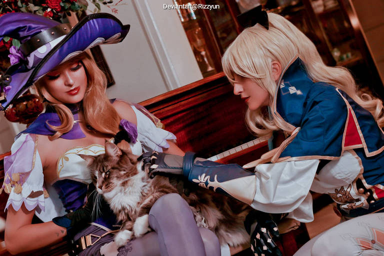 Two individuals in fantasy-themed costumes are interacting with a fluffy cat, with one person wearing a purple hat and the other dressed in a blue and white outfit.