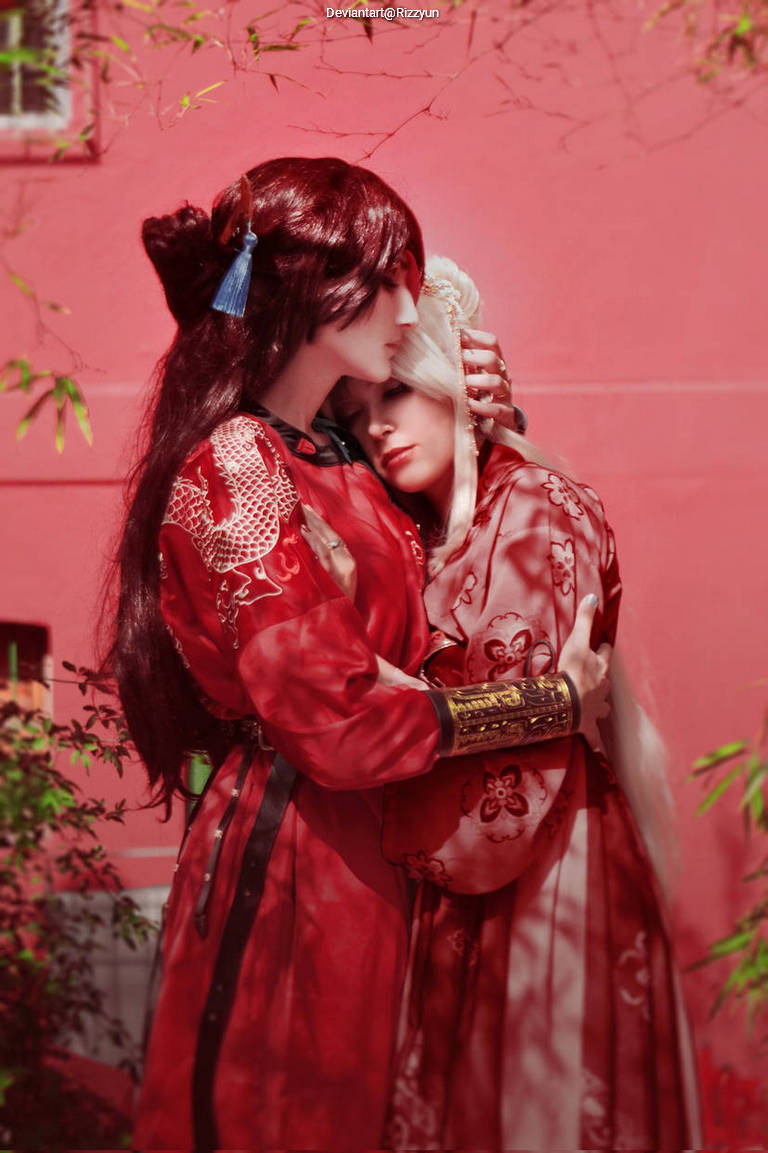 Two individuals in red traditional attire embrace each other against a soft pink background, depicting a moment of affection and intimacy.