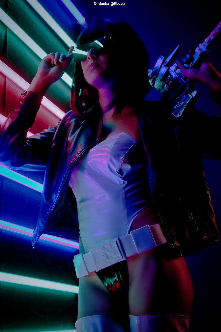 A person in a futuristic outfit with a leather jacket and neon-colored lights in the background, holding a pair of glasses and posing confidently.