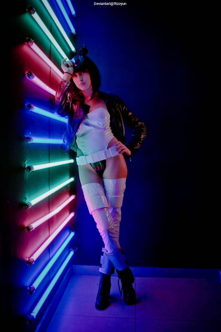 A person with short dark hair is posing against a wall illuminated by colorful neon lights. They are wearing a black leather jacket and white attire, creating a bold fashion statement.