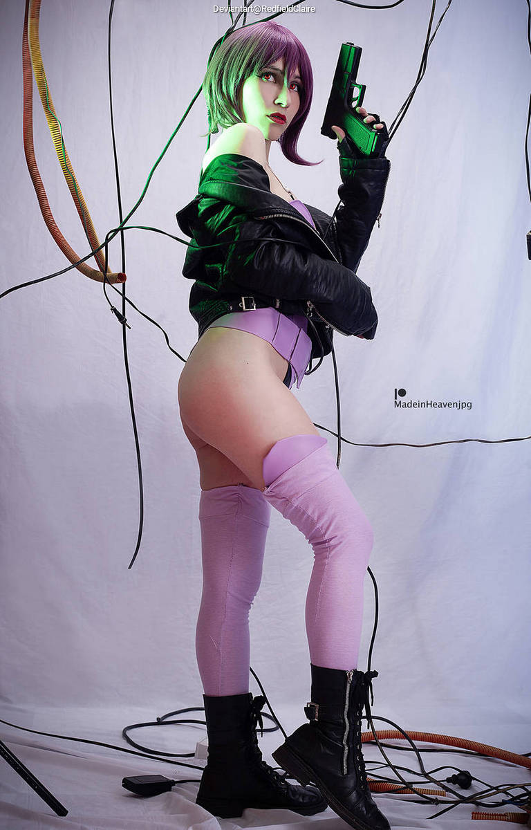 A cosplayer poses with a gun while wearing a black leather jacket, purple bodysuit, and thigh-high stockings, standing against a backdrop of wires and a white sheet.