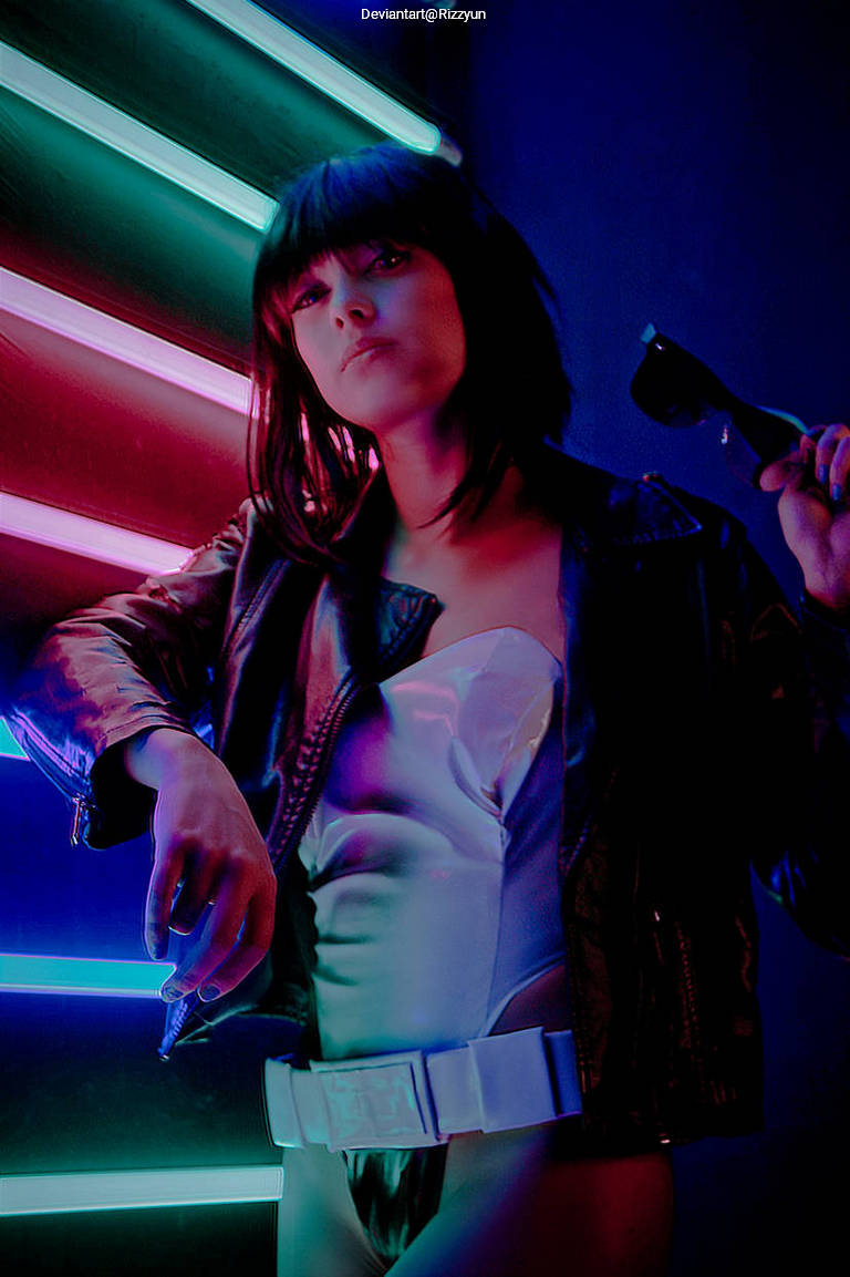 A person with short dark hair poses in front of neon lights, wearing a shiny outfit and a leather jacket, while holding sunglasses.