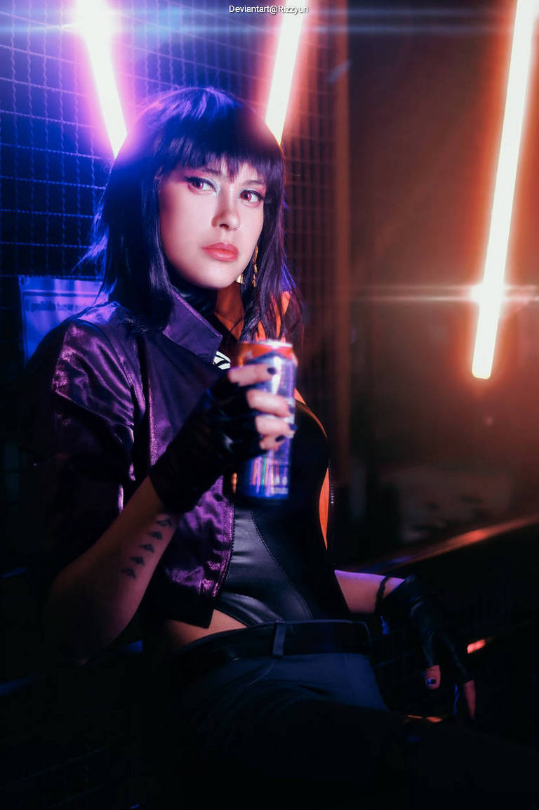 A woman in a purple jacket poses confidently, holding a can, with vibrant neon lights in the background.