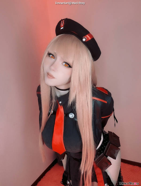 A person in a stylish costume with a black and red uniform, long blonde hair, and striking orange eyes, posing playfully against a softly lit pink background.