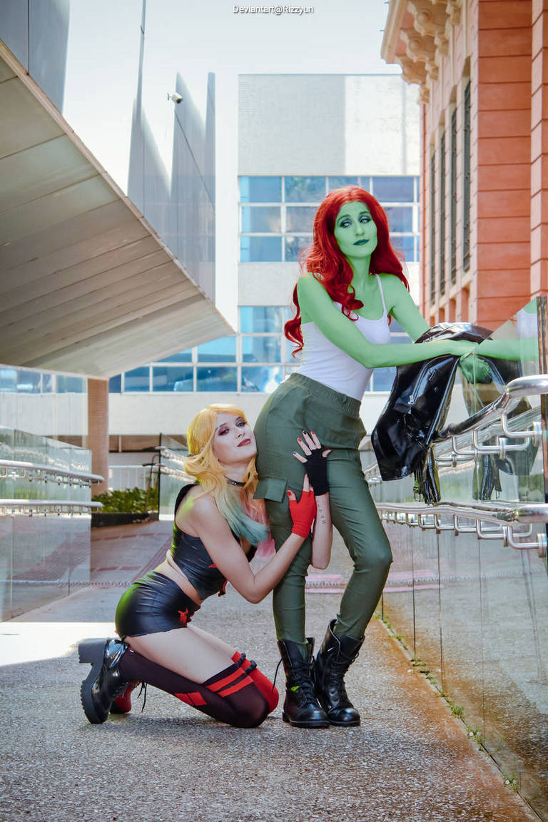 Two individuals in costume as comic book characters, one with green skin and red hair, standing confidently, and the other kneeling beside her, dressed in a black and red outfit.