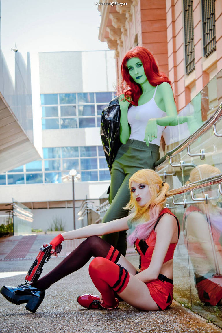 Two women in vibrant costumes posing outdoors. One has green skin and red hair, wearing a tank top and skirt. The other has blonde hair with pink highlights, dressed in a red outfit with stockings and holding a prop gun.