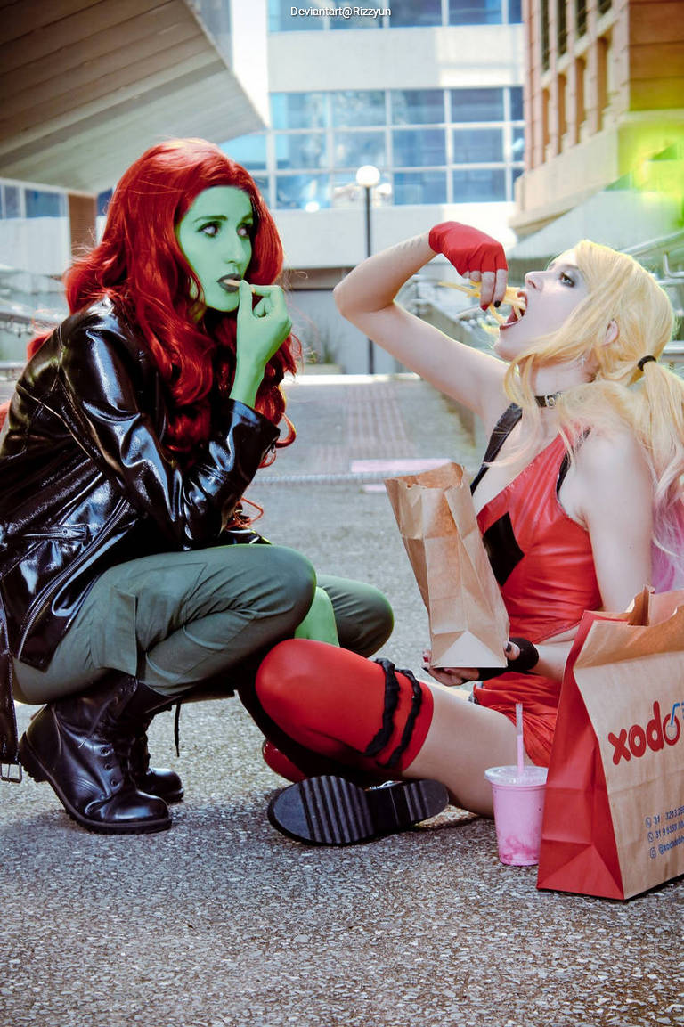 Two women in cosplay, one with green skin and red hair eating, and the other with blonde hair in red and black attire, enjoying food together outdoors.
