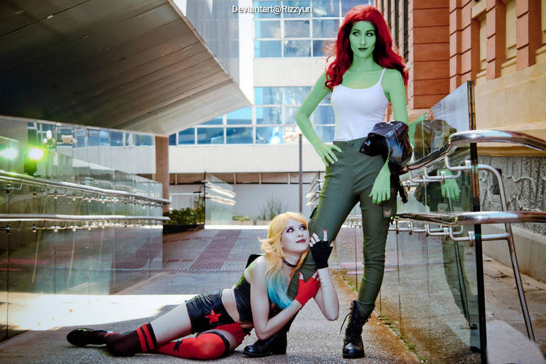 A female cosplayer portraying Harley Quinn in a playful pose and another cosplayer as Poison Ivy, standing confidently in an urban setting with modern architecture in the background.