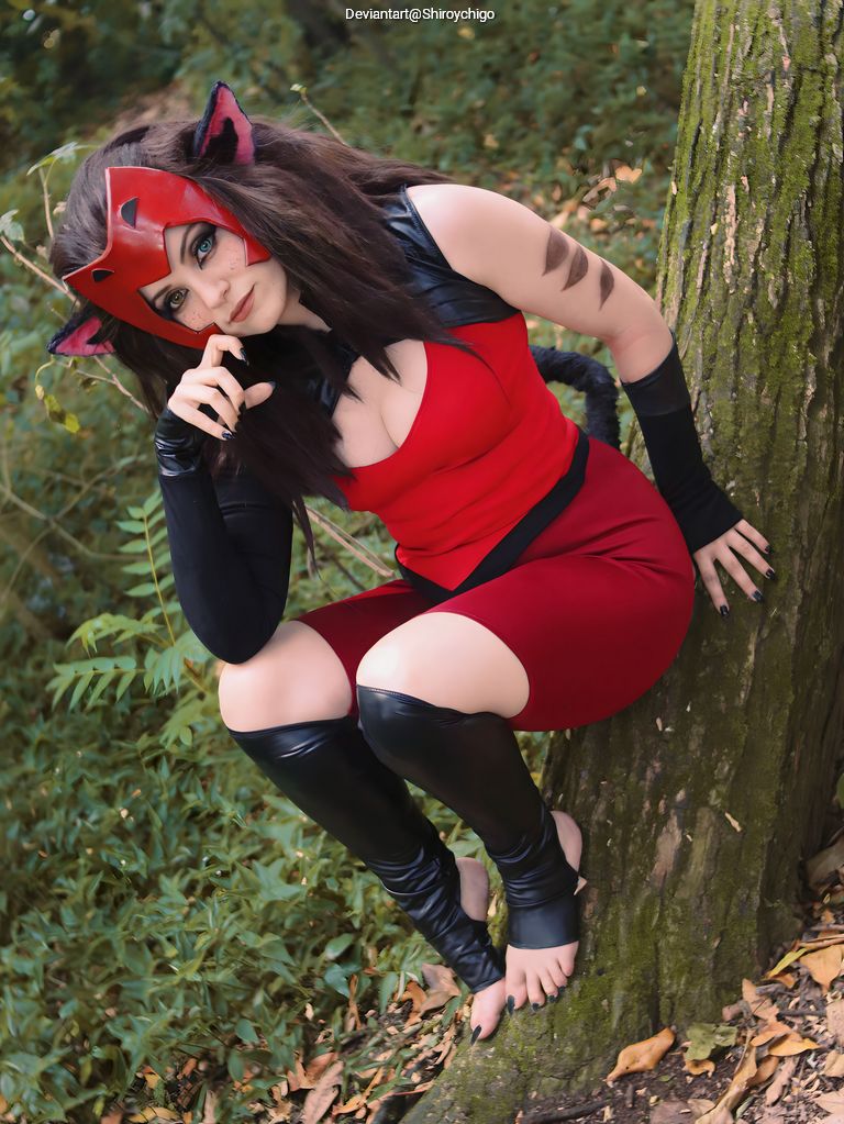 A person dressed in a red and black costume with cat-like features, including cat ears and claws, sitting against a tree in a forested area.