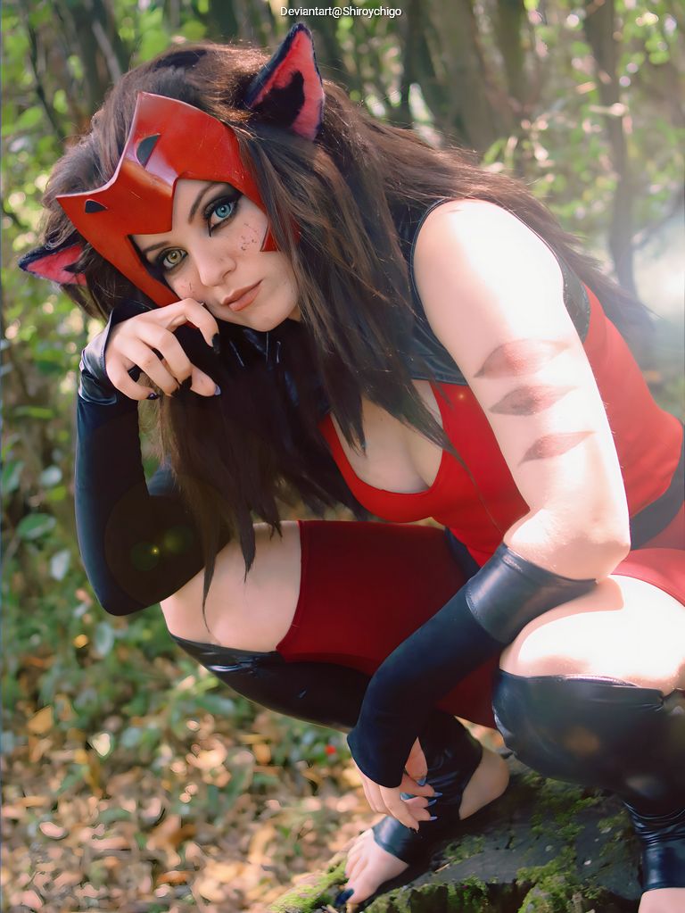 A person cosplaying as a character with a red costume and mask, featuring cat ears, crouching on a rock in a forest setting, with a thoughtful pose.