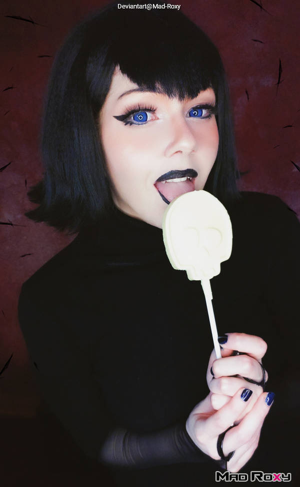 A person with short black hair and striking blue eyes poses with a skull-shaped lollipop, wearing dark makeup and a black outfit against a red backdrop.