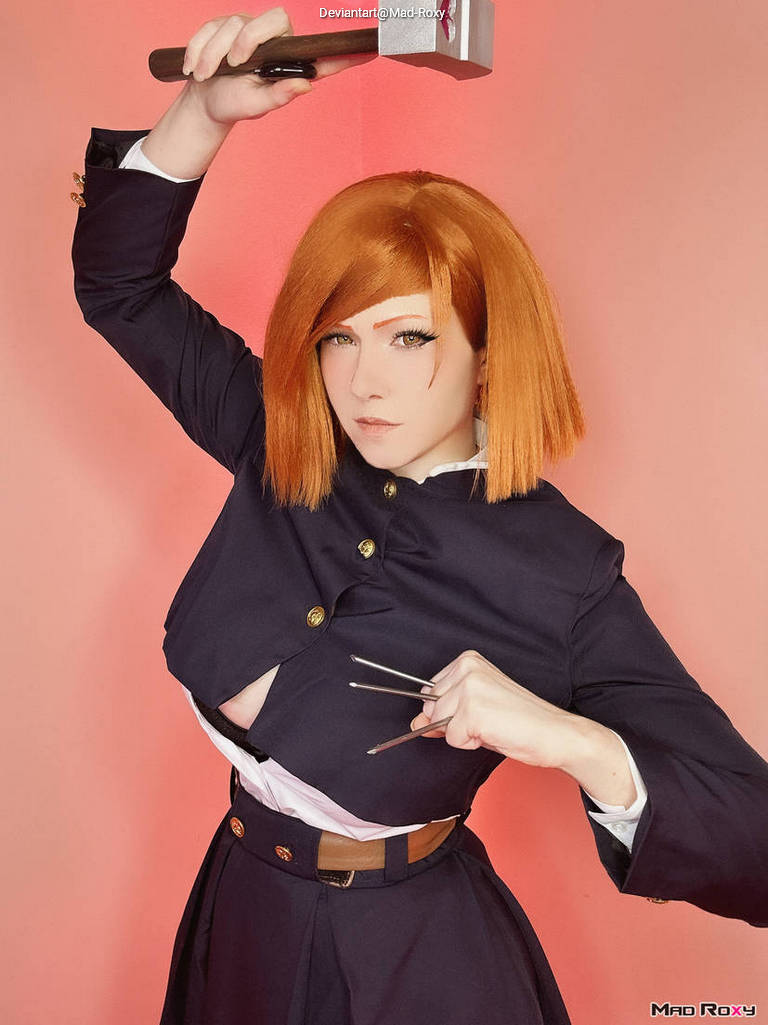 A person in a dark outfit striking a fierce pose while holding a hammer in one hand and displaying claws with the other, set against a pink background.