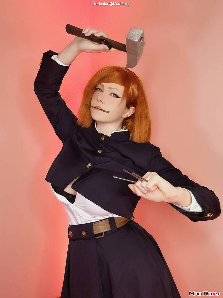 A person in a stylish, dark outfit holds a hammer above their head while displaying a playful expression. They have short orange hair and are posed against a pink background.