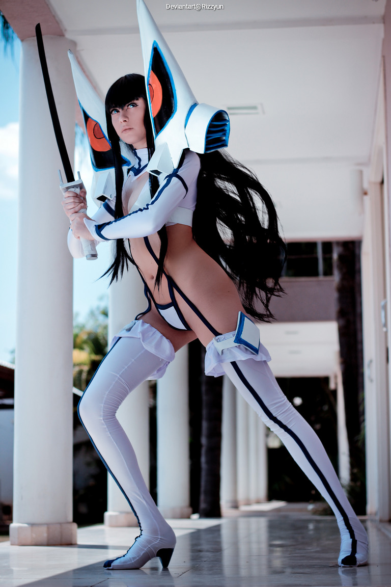 A female cosplayer dressed in a futuristic outfit, wielding a sword, with long dark hair and distinctive accessories, standing in an outdoor corridor.