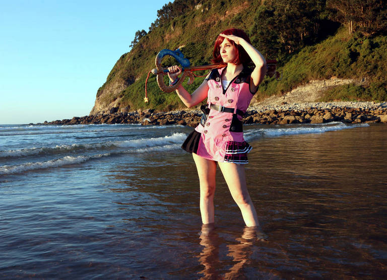 A person in a pink and black costume stands in shallow water at the beach, shielding their eyes from the sun while holding a colorful staff. A rocky shore and a green hillside are visible in the background.