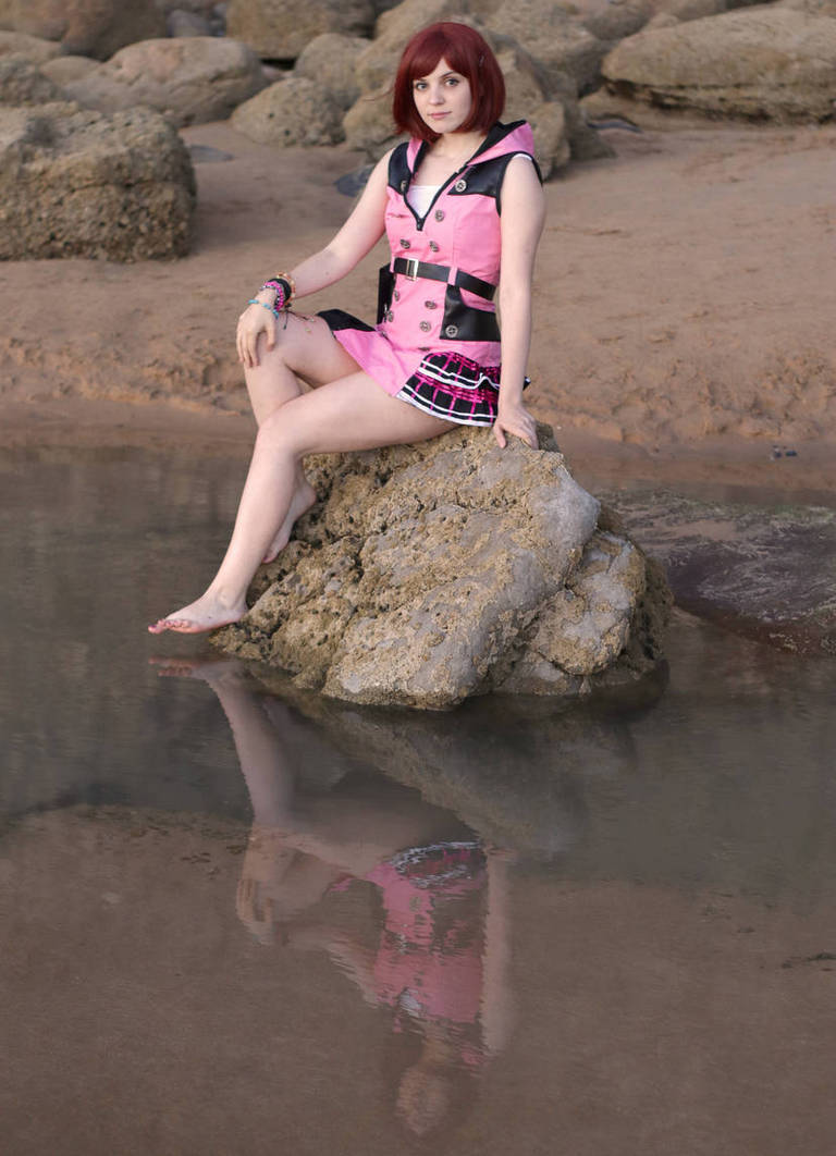A person sitting on a rock by the water's edge, wearing a pink and black outfit, with short red hair, reflecting in the water.