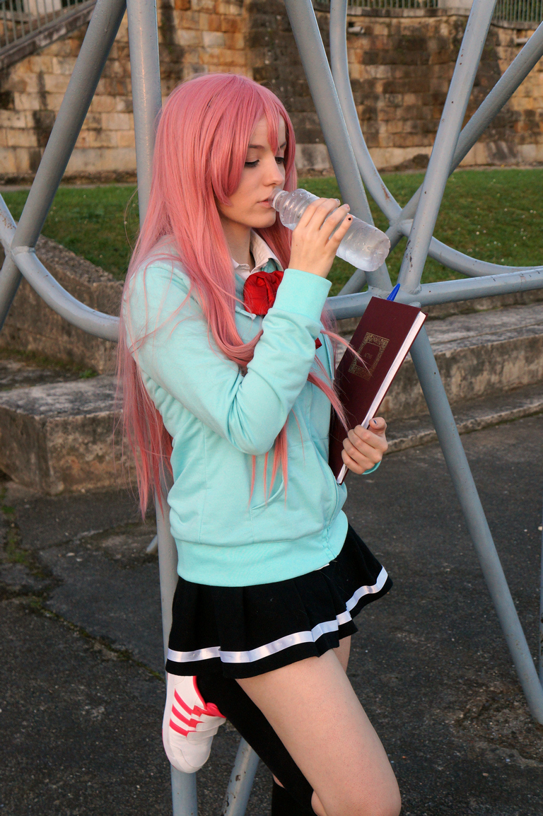 Young person with long pink hair, wearing a light blue hoodie and a black skirt, drinking from a plastic water bottle while holding a book.