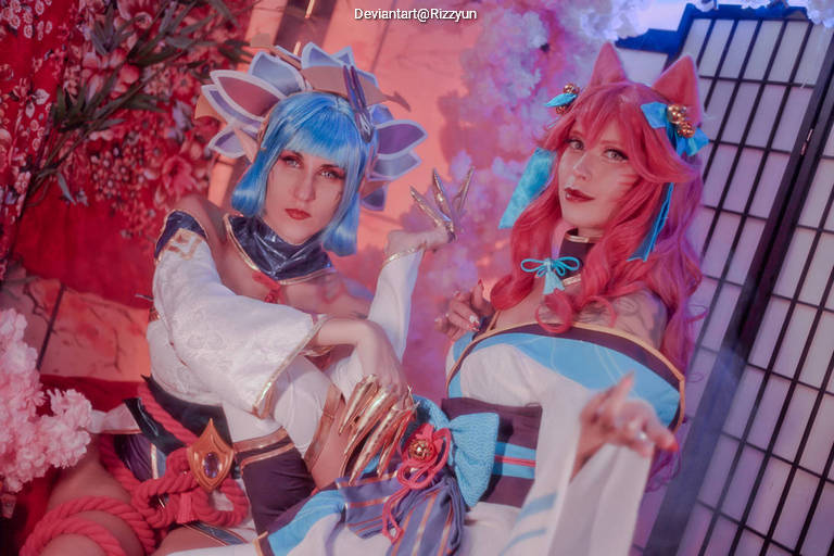 Two individuals in elaborate cosplay costumes, one with blue hair styled in a floral theme and the other with red hair featuring cat ears, posing in front of a backdrop decorated with cherry blossoms and traditional Japanese elements.