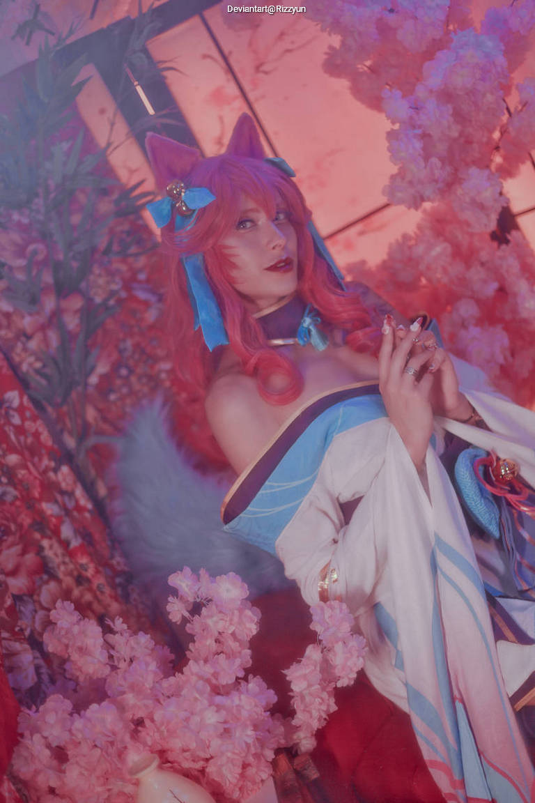 A character with pink hair and fox-like ears, dressed in a blue and white outfit, poses among pink cherry blossoms in a beautifully lit setting.