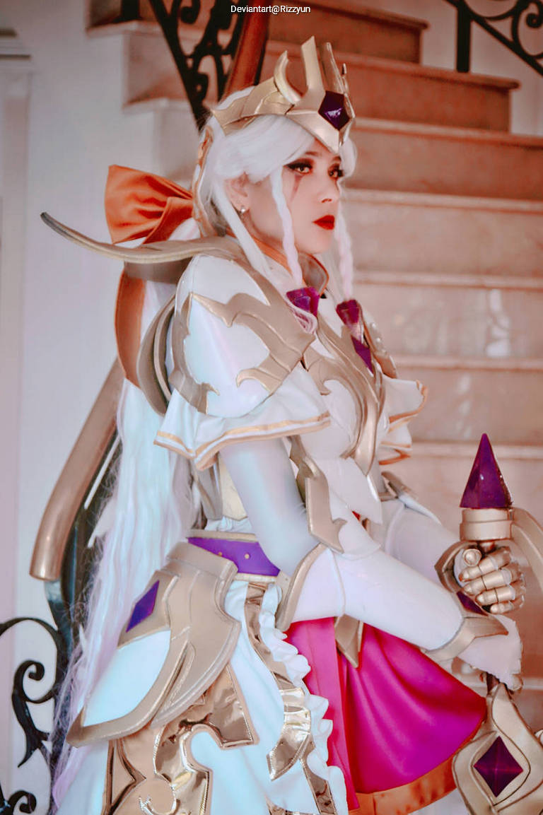 A person in elaborate fantasy cosplay, featuring a white and gold armor with purple accents, a long white wig with braids, and a colorful bow. The background includes a staircase with decorative railings.