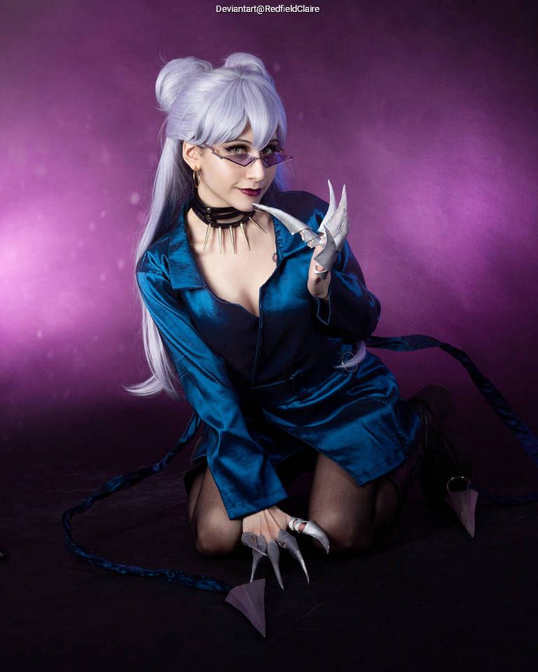 A person with long silver hair wearing blue clothing and accessories, posing on the floor with a playful expression. The background features a purple gradient.