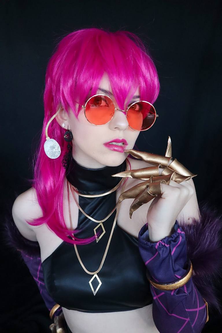 A person with bright pink hair and sunglasses poses confidently, wearing a black outfit with gold accessories and a long-sleeved purple garment.