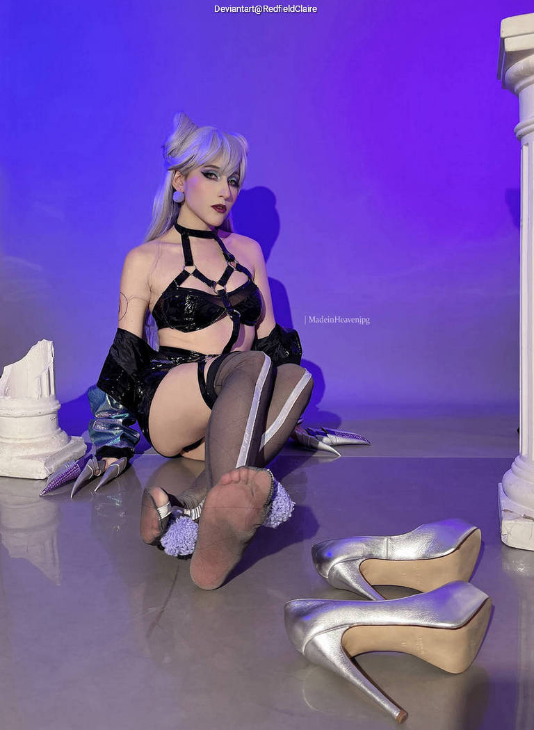 A person sitting on the floor in a costume that features a black outfit with sheer elements, silver high heels nearby, and a purple backdrop.