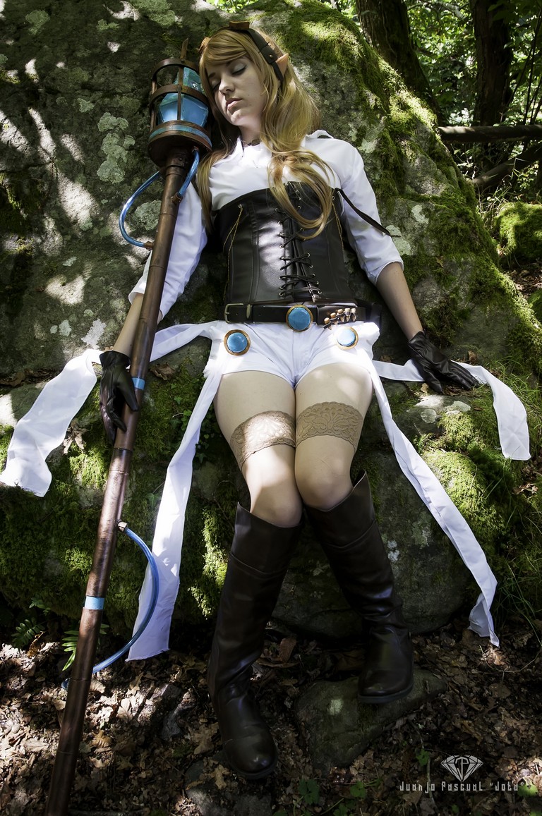 A woman in a fantasy-themed costume, sitting against a mossy rock in a forest, holding a staff. She has long blonde hair and wears a corset, shorts, and thigh-high boots.