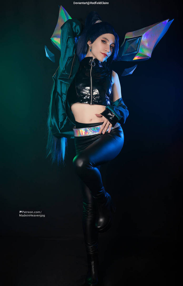 A person posing confidently in a stylish black and teal costume with shiny accents, featuring a crop top and high-waisted pants. The background is dimly lit with colorful lights.