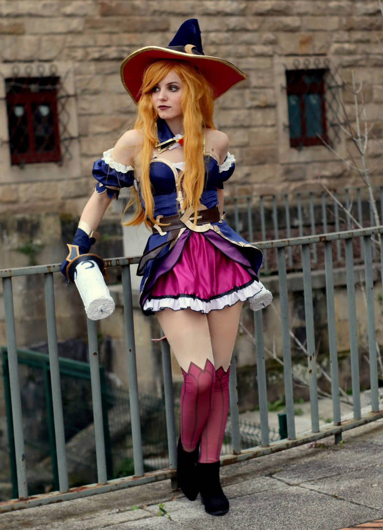 A person in a colorful wizard costume with a large hat, standing by a railing in front of a stone wall. The outfit features a purple and pink skirt, white lace trim, and long, flowing orange hair.