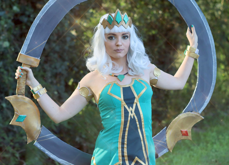 A person in a vibrant teal costume with gold accents, styled hair, and a jeweled crown, posed confidently while holding a large circular weapon.