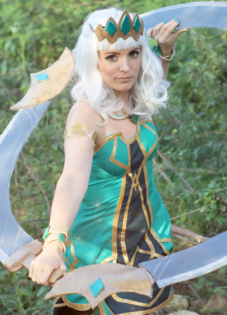 A person in a green and gold costume, resembling a fantasy character, wielding large curved weapons in a nature setting.