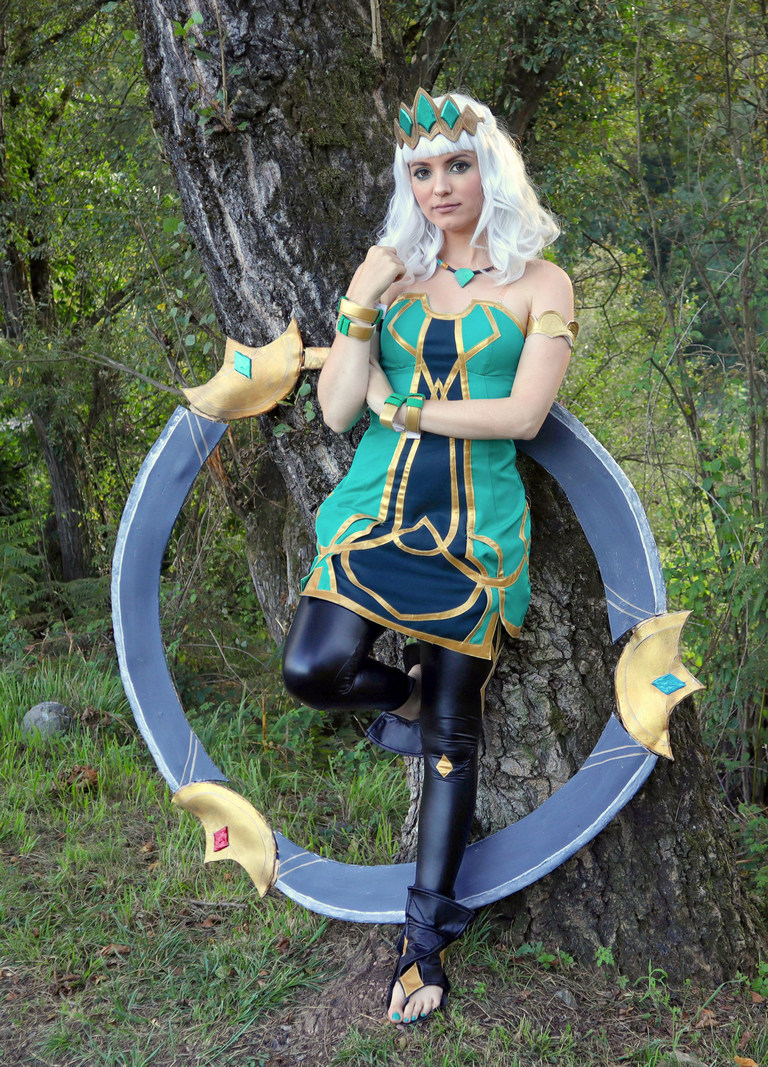 A woman wearing a green and gold costume with a crown, standing next to a large circular prop. She has long white hair and is posed against a tree in a natural outdoor setting.
