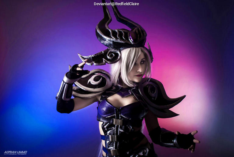 A female cosplayer dressed as a character with elaborate dark armor and a distinct headpiece. The background features vibrant purple and blue lighting.