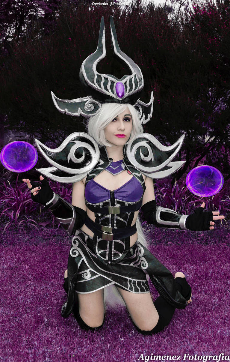 A person in elaborate cosplay, featuring a dark-themed outfit with swirling patterns and a purple gemstone, kneels in a vibrant purple setting, holding glowing orbs.